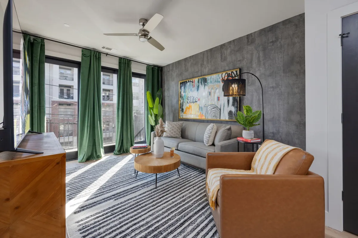 A stylish living room bathed in sunlight, featuring a plush sofa and accent chair arranged around a modern coffee table. Lush green curtains frame the windows, vibrant artwork adds a creative touch, and the inviting atmosphere suggests a perfect space for relaxing, reading, or entertaining guests.