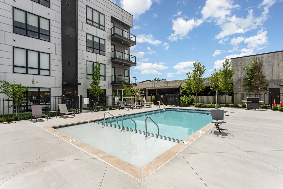 A tranquil exterior shot showcases the year-round swimming pool surrounded by lush greenery and building balconies. The poolside area features inviting lounge seating and hints of communal spaces for social gatherings or quiet afternoons.