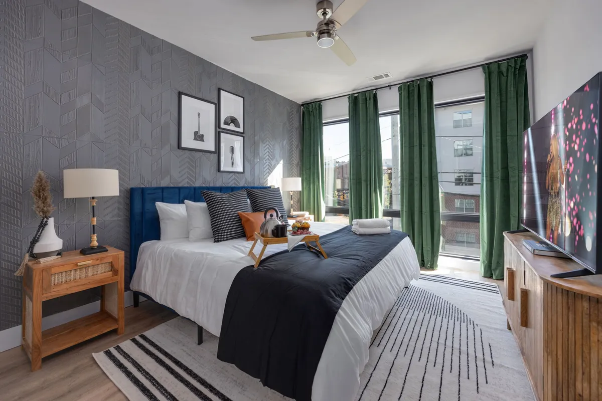 A stylish bedroom with navy and white bedding, large windows with green curtains that let in ample natural light, a TV on a wooden media console, and decorative wall art. The setting feels cozy and serene, ideal for restful nights or lazy mornings.