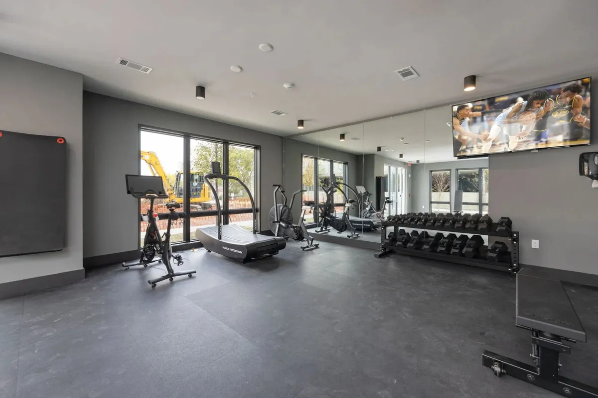 A bright, modern fitness center filled with natural light from large windows. The room includes a treadmill, dumbbell rack, exercise bike, and a TV screen—supporting both personal and group fitness activities.