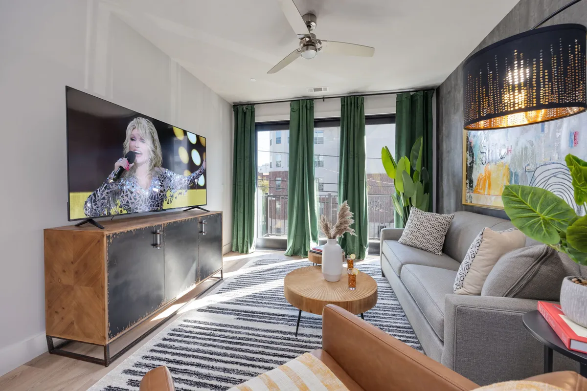 A vibrant living room scene with a modern sofa, large TV set for entertainment, green curtains framing the windows, a wooden coffee table, and decorative wall art. The space exudes energy and comfort, ideal for both relaxed evenings and social gatherings.