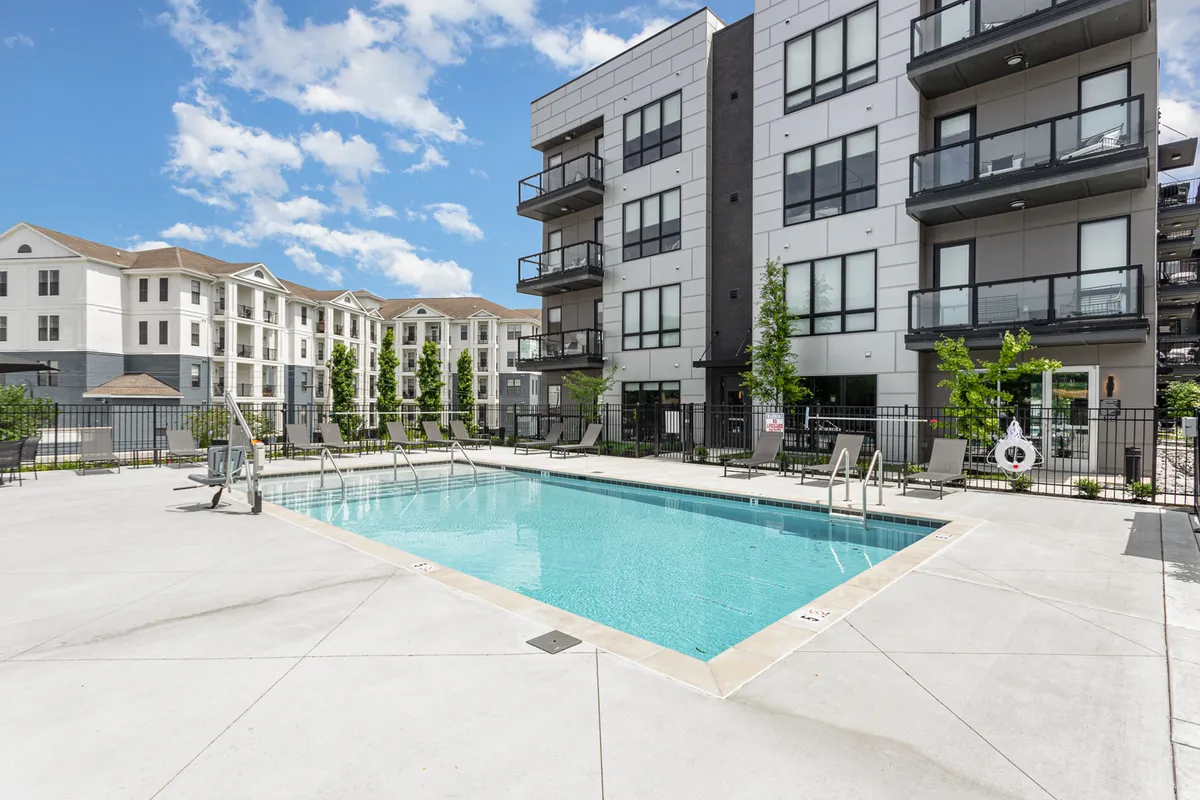 This image features a lively exterior pool area surrounded by balconies and lush green landscaping. People are seen swimming, sunbathing, socializing, and relaxing by the water, capturing the vibrant, resort-style atmosphere.
